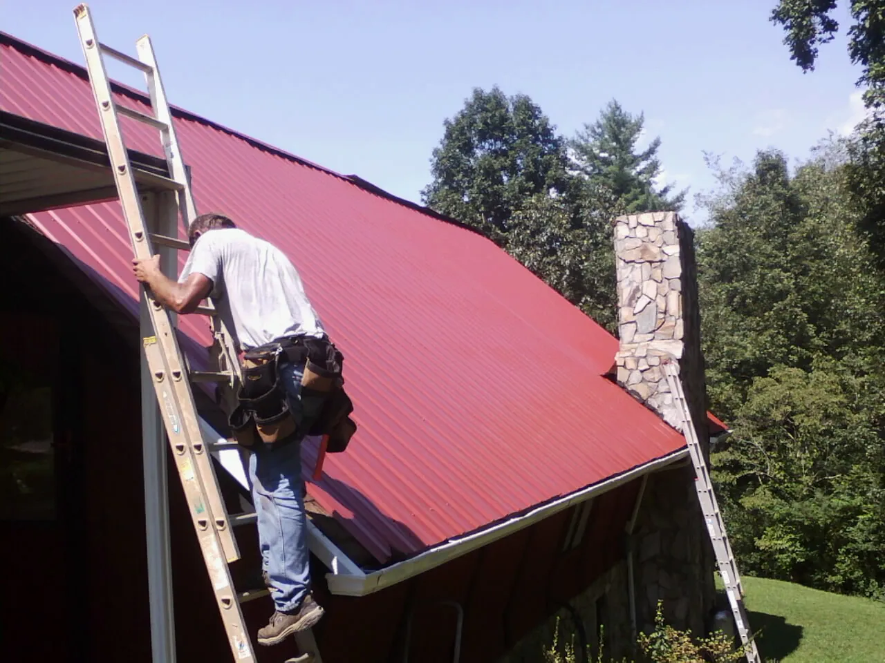 Roofer on a ladder beside a red metal roof and stone chimney — Soffit & Fascia Repair by Roofing Brothers in Buckeystown